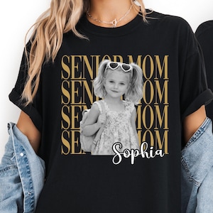 Pu&ograve; includere: Una t-shirt nera a maniche corte con una grafica dorata ripetuta che recita SENIOR MOM. Al centro &egrave; presente una fotografia in bianco e nero di una bambina con le codine, occhiali da sole a forma di cuore e uno zaino, con il nome Sophia.