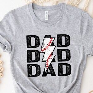 May include: A light gray t-shirt with the word "DAD" stacked three times in a bold, distressed black font. A white lightning bolt with a baseball design in the center replaces the letter "D". The baseball has red stitching.