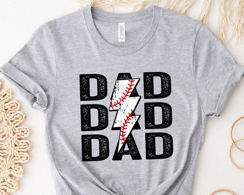 May include: A light gray t-shirt with the word "DAD" stacked three times in a bold, distressed black font. A white lightning bolt with a baseball design in the center replaces the letter "D". The baseball has red stitching.