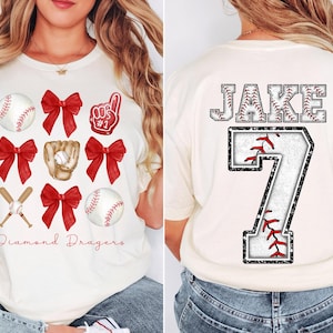 May include: White t-shirt with red and white baseball themed graphics. The front of the shirt has a baseball, bats, bows, and a glove. The back of the shirt has the name "JAKE" and the number "7" in a baseball-themed font.