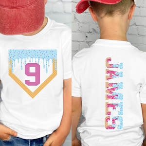 May include: White t-shirt with a baseball home plate design featuring a number 9 and blue icing drips. The back of the shirt displays the name "JAMES" vertically in a donut-themed font. The shirt is worn by a person wearing a red baseball cap.