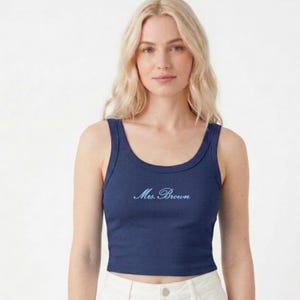 May include: A navy blue ribbed tank top with a scoop neck. The text "Mrs. Brown" is embroidered in a light blue script across the chest. The top is a cropped style.
