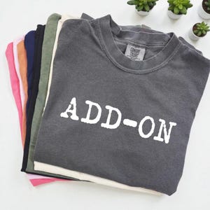 May include: A stack of various colored t-shirts, including pink, orange, navy, and gray. The top shirt is gray with the white text "ADD-ON" printed on the front. The shirts are neatly folded and stacked.