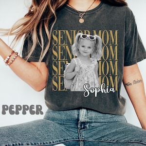 Pu&ograve; includere: Una t-shirt a maniche corte grigio antracite con il testo dorato ripetuto SENIOR MOM e una fotografia in bianco e nero di una bambina con le codine che indossa occhiali da sole a forma di cuore e uno zaino, con il nome Sophia scritto sotto.