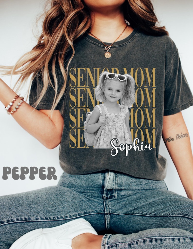 Pu&ograve; includere: Una t-shirt a maniche corte grigio antracite con il testo dorato ripetuto SENIOR MOM e una fotografia in bianco e nero di una bambina con le codine che indossa occhiali da sole a forma di cuore e uno zaino, con il nome Sophia scritto sotto.