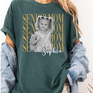 Pu&ograve; includere: Una t-shirt verde scuro con una grafica dorata ripetuta che recita SENIOR MOM. Al centro c'&egrave; una fotografia in bianco e nero di una bambina con le codine, occhiali da sole a forma di cuore e uno zaino, con il nome Sophia in corsivo bianco.