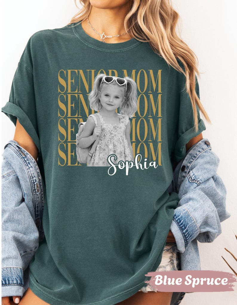 Pu&ograve; includere: Una t-shirt verde scuro con una grafica dorata ripetuta che recita SENIOR MOM. Al centro c'&egrave; una fotografia in bianco e nero di una bambina con le codine, occhiali da sole a forma di cuore e uno zaino, con il nome Sophia in corsivo bianco.