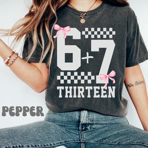 May include: A charcoal gray short-sleeve t-shirt featuring a white graphic print of the numbers 6 and 7 separated by a plus sign, with the word THIRTEEN below. The design includes two pink bows and black and white checkered patterns.