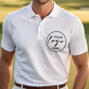 May include: White polo shirt with a collar and short sleeves. The shirt features a three-button placket and a circular logo design on the chest that says "YOUR logo HERE" in black. The model is wearing a brown belt.