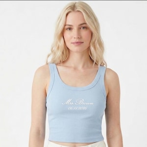 May include: A light blue ribbed tank top with a scoop neck. The text "Mrs. Brown 06.11.2026" is embroidered in white script across the chest. The top is worn by a person with blonde hair.