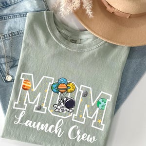 Puede incluir: Una camiseta verde salvia con las palabras "MOM Launch Crew" en letras blancas. El dise&ntilde;o presenta un astronauta flotando con globos de planetas. La camiseta est&aacute; doblada sobre unos vaqueros azules y un sombrero beige.