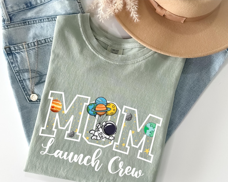 Puede incluir: Una camiseta verde salvia con las palabras "MOM Launch Crew" en letras blancas. El dise&ntilde;o presenta un astronauta flotando con globos de planetas. La camiseta est&aacute; doblada sobre unos vaqueros azules y un sombrero beige.