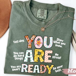 May include: Moss green t-shirt with the words "YOU ARE READY" in colorful, patterned letters. The shirt includes phrases like "Test day" and "Do your best." A gold necklace with a pendant rests on the shirt.