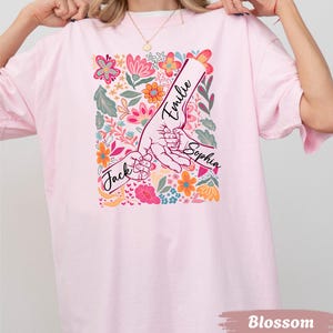 Op de afbeelding: Lichtroze t-shirt met een bloemenontwerp en de namen "Jack", "Emilie" en "Sophia" erop gedrukt. Het ontwerp toont handen die elkaar vasthouden, omringd door kleurrijke bloemen. Het woord "Blossom" staat onderaan.