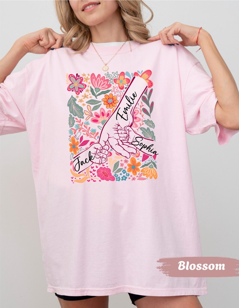 Op de afbeelding: Lichtroze t-shirt met een bloemenontwerp en de namen "Jack", "Emilie" en "Sophia" erop gedrukt. Het ontwerp toont handen die elkaar vasthouden, omringd door kleurrijke bloemen. Het woord "Blossom" staat onderaan.