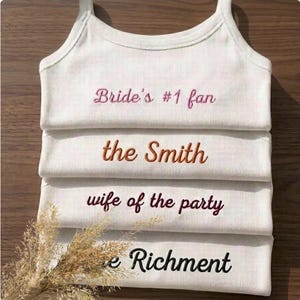 May include: White tank tops with embroidered text. The text includes "Bride's #1 fan", "the Smith", "wife of the party", and "e Richment". The tank tops are stacked on top of each other.
