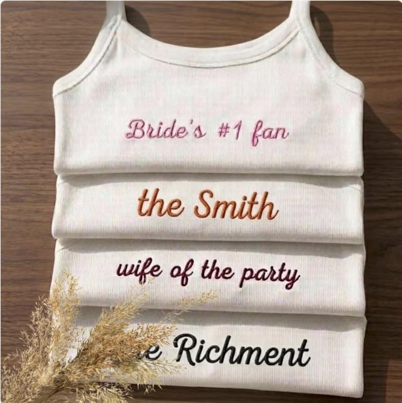 May include: White tank tops with embroidered text. The text includes "Bride's #1 fan", "the Smith", "wife of the party", and "e Richment". The tank tops are stacked on top of each other.
