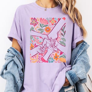 Op de afbeelding: Een lichtpaars T-shirt met een bloemenontwerp en handen die elkaar vasthouden. De afbeelding bevat kleurrijke bloemen in roze, oranje en blauw. Het shirt is gemaakt van een zacht materiaal.
