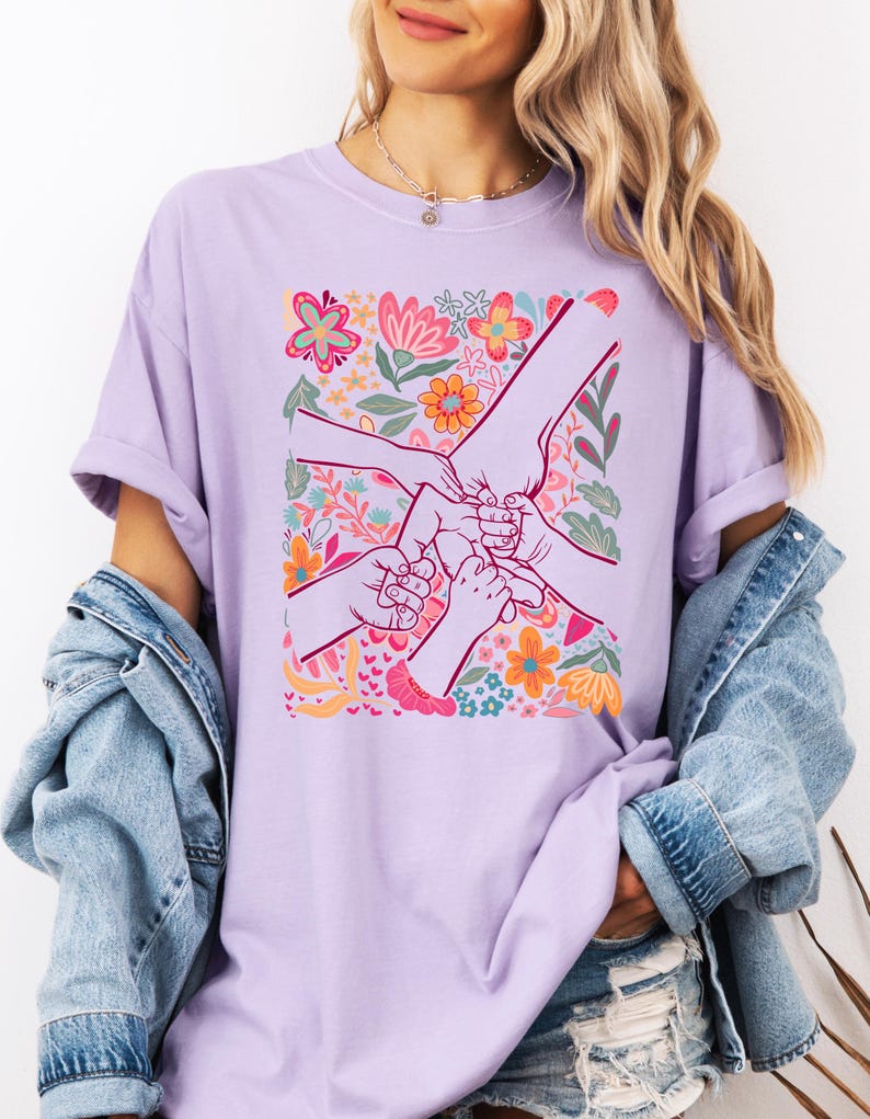 Op de afbeelding: Een lichtpaars T-shirt met een bloemenontwerp en handen die elkaar vasthouden. De afbeelding bevat kleurrijke bloemen in roze, oranje en blauw. Het shirt is gemaakt van een zacht materiaal.