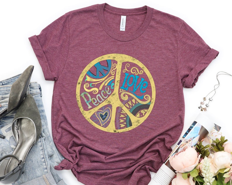 Op de afbeelding: Een maroon t-shirt met een peace-teken graphic. Het peace-teken is geel met een blauw en groen ontwerp en de woorden "Peace" en "Love" erin.