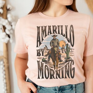 May include: A pink t-shirt with a vintage graphic design featuring a cowboy riding a horse with the text "Amarillo by Morning".