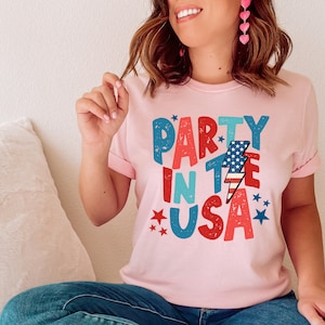 May include: A pink t-shirt with the text "Party in the USA" in colorful, distressed lettering. The text is decorated with stars and a lightning bolt with an American flag design.