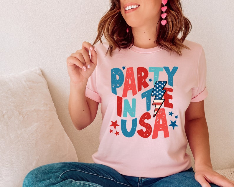 May include: A pink t-shirt with the text "Party in the USA" in colorful, distressed lettering. The text is decorated with stars and a lightning bolt with an American flag design.