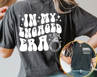 Könnte beinhalten: Ein schwarzes T-Shirt mit weißem Text, der "In My Engaged Era" lautet, mit einer stilisierten Ring- und Diamantgrafik. Die Rückseite des Shirts hat weißen Text, der "Future Mrs. Marshall" lautet.