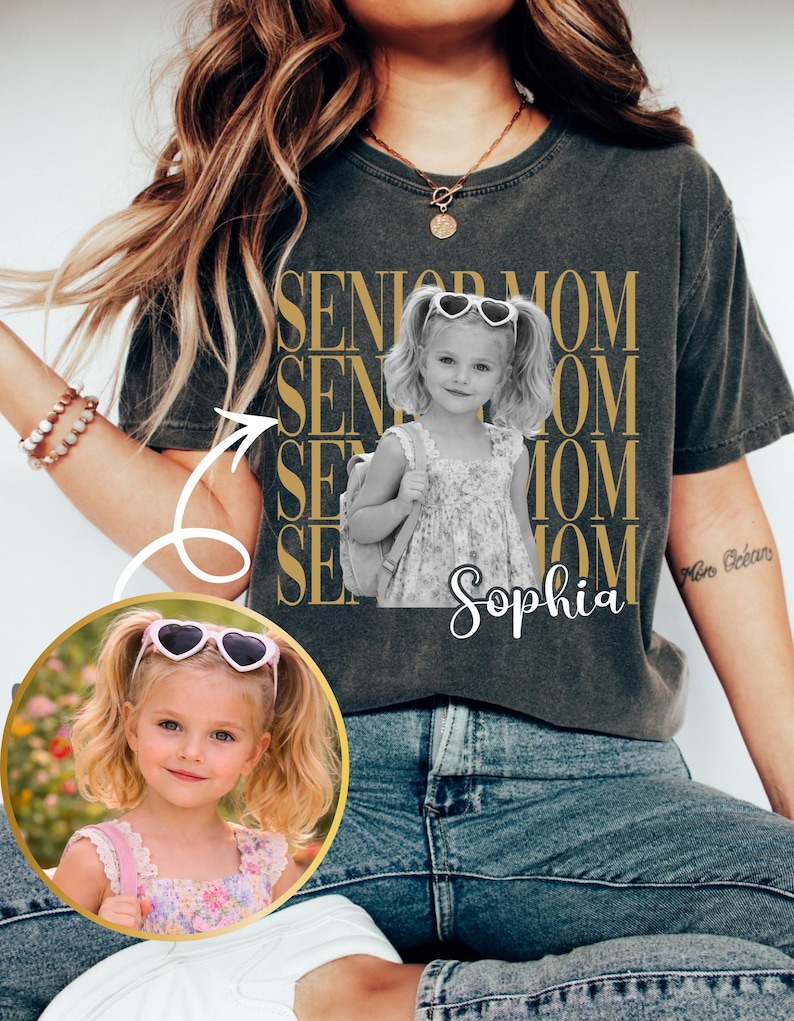 Pu&ograve; includere: Una t-shirt grigio antracite con il testo dorato ripetuto SENIOR MOM e una fotografia in bianco e nero di una bambina con le codine che indossa occhiali da sole a forma di cuore e uno zaino, con il nome Sophia scritto in bianco sotto.