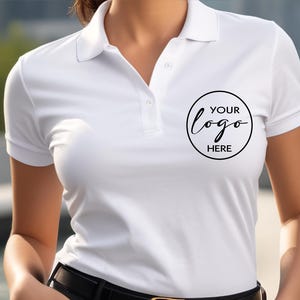 May include: White polo shirt with short sleeves and a collar. The shirt has a black circle with the text "YOUR logo HERE" printed on the chest. The shirt features a button-down placket and is made of a smooth fabric.