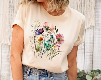 Könnte beinhalten: Ein cremefarbenes Kurzarm-T-Shirt mit einer lebendigen Aquarellgrafik verschiedener Wildblumen in Lila-, Rosa-, Orange-, Blau- und Grüntönen. Das T-Shirt hat einen Rundhalsausschnitt und wird mit einer blauen Jeans kombiniert.