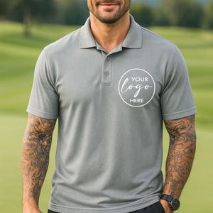 May include: Grey polo shirt with a three-button placket and short sleeves. The shirt features a white circular logo with the text "YOUR logo HERE". The model is wearing a black watch.