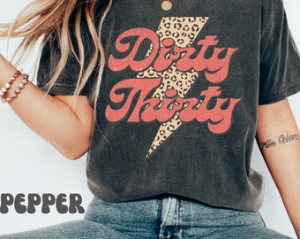 May include: A dark gray t-shirt with a red and white graphic that says "Dirty Thirty" with a leopard print lightning bolt. The shirt is distressed and has a vintage look.