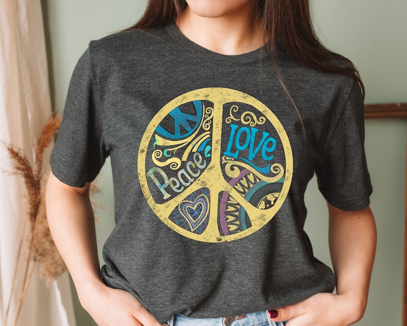 Op de afbeelding: Een donkergrijs T-shirt met een gele peace-teken graphic. Het peace-teken is versierd met blauwe, paarse en groene swirls en de woorden "Peace" en "Love" zijn in het teken geschreven.