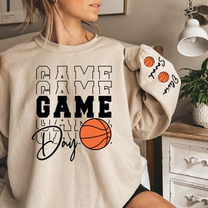 May include: A beige sweatshirt with the text "GAME DAY" in black letters and a basketball graphic. The sleeves have basketball graphics and the text "Senior" and "Junior" in black.