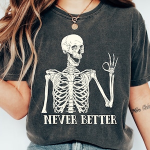 May include: A dark gray t-shirt with a white skeleton graphic giving the OK sign. The text "NEVER BETTER" is printed below the skeleton.