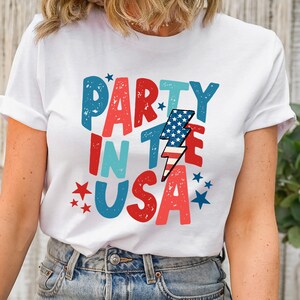 May include: White t-shirt with a red, white, and blue graphic that says "Party in the USA" with a lightning bolt and stars.