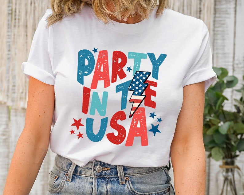 May include: White t-shirt with a red, white, and blue graphic that says "Party in the USA" with a lightning bolt and stars.