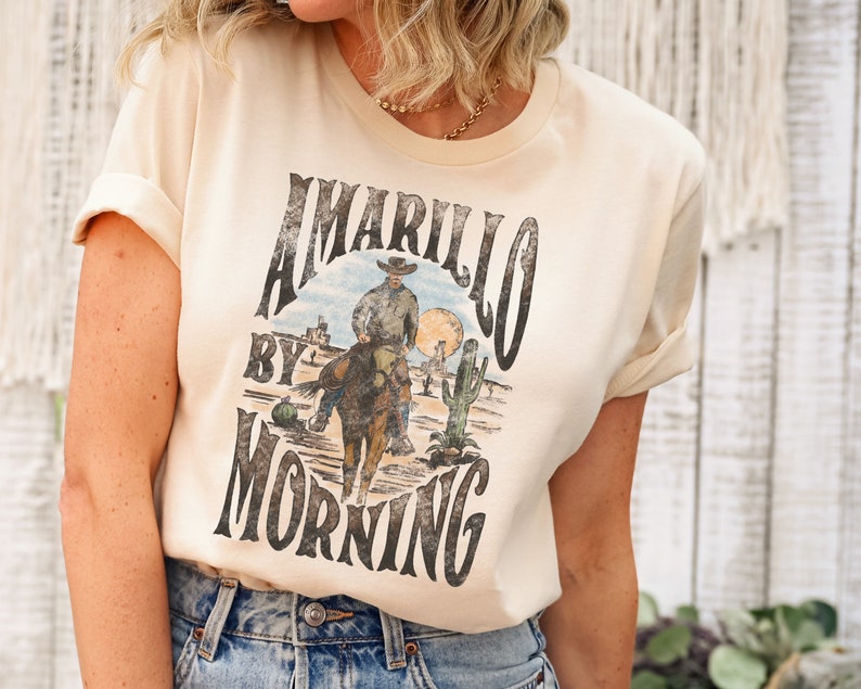 May include: A cream coloured t-shirt with a vintage graphic design featuring a cowboy riding a horse in a desert landscape. The text on the shirt reads "Amarillo by Morning".