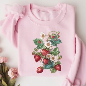 Pode incluir: Uma camisa de moletom rosa com uma ilustra&ccedil;&atilde;o vintage de uma planta de morango com morangos vermelhos e flores brancas.