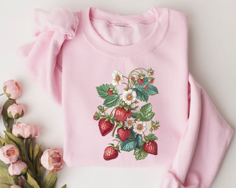 Pode incluir: Uma camisa de moletom rosa com uma ilustra&ccedil;&atilde;o vintage de uma planta de morango com morangos vermelhos e flores brancas.