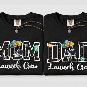 Puede incluir: Dos camisetas negras con las palabras "MOM Launch Crew" y "DAD Launch Crew" en blanco. Cada camiseta presenta un gr&aacute;fico tem&aacute;tico espacial con planetas, un astronauta y la etiqueta Comfort Colors.