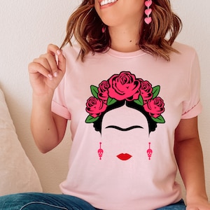 May include: A pink t-shirt with a black and white illustration of Frida Kahlo wearing a flower crown. The crown is made of red roses with green leaves. The illustration also includes Frida Kahlo's signature unibrow and mustache.