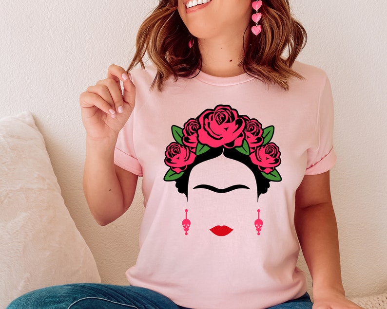 May include: A pink t-shirt with a black and white illustration of Frida Kahlo wearing a flower crown. The crown is made of red roses with green leaves. The illustration also includes Frida Kahlo's signature unibrow and mustache.