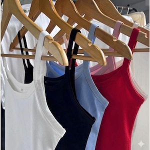 May include: A row of colorful tank tops hanging on wooden hangers. The tank tops are in various colors, including white, black, light blue, and red. The tops have thin straps and a simple, classic design, perfect for layering or warm weather.