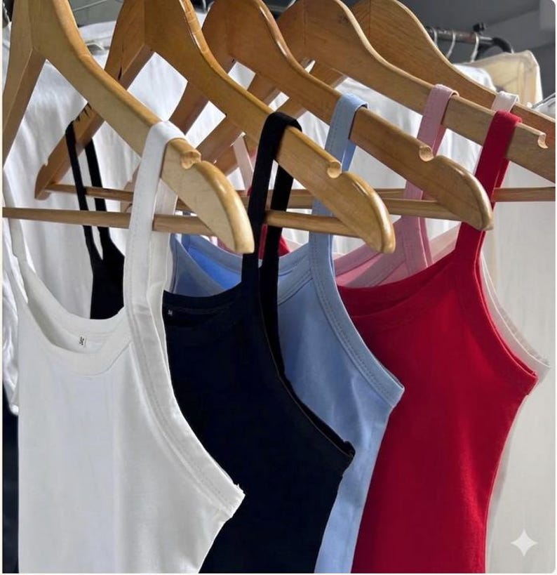 May include: A row of colorful tank tops hanging on wooden hangers. The tank tops are in various colors, including white, black, light blue, and red. The tops have thin straps and a simple, classic design, perfect for layering or warm weather.