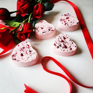 Könnte beinhalten: Vier herzförmige, rosa Badebomben, bestreut mit roten Rosenblättern. Ein Strauß roter Rosen und ein rotes Band sind auf einer weißen Oberfläche angeordnet, was ein romantisches Thema ergibt. Die Badebomben haben eine hellrosa Farbe.