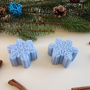 May include: Two light blue snowflake-shaped soaps with a raised snowflake design. The soaps are on a white surface with cinnamon sticks and a star anise.