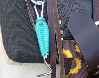 May include: A close-up of a turquoise paracord wrapped horse grooming tool attached to a saddle with brown leather straps and a sunflower patterned strap.