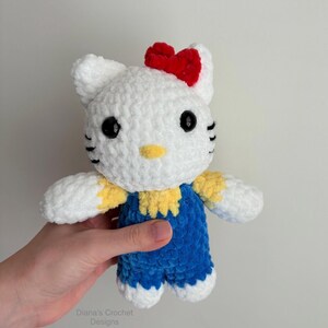 May include: A handmade white crochet cat doll with a red bow, black eyes, and yellow accents. The cat wears blue overalls. The doll is approximately 6 inches tall and is a cute, soft toy.